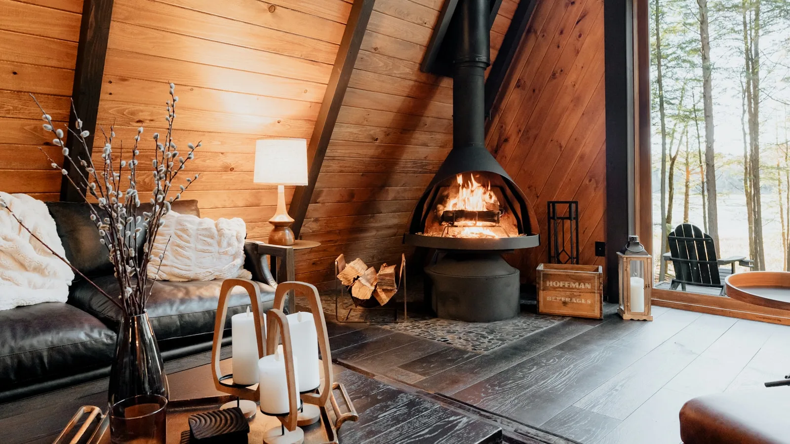 Interior cálido de una cabaña con chimenea de leña encendida, sofá de cuero, velas y vistas al bosque.