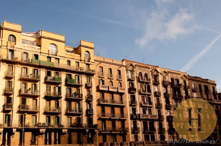 Fachada de edificios residenciales en una calle urbana bajo cielo azul con nubes dispersas y un logo circular superpuesto
