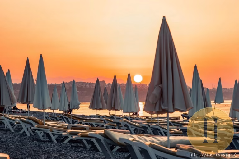 Playa con tumbonas y sombrillas cerradas al atardecer, con el sol bajo y una ciudad al fondo