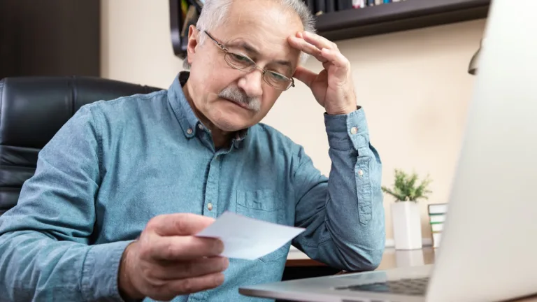 Hombre mayor con gafas y camisa azul, mirando un papel con expresión preocupada frente a un portátil.
