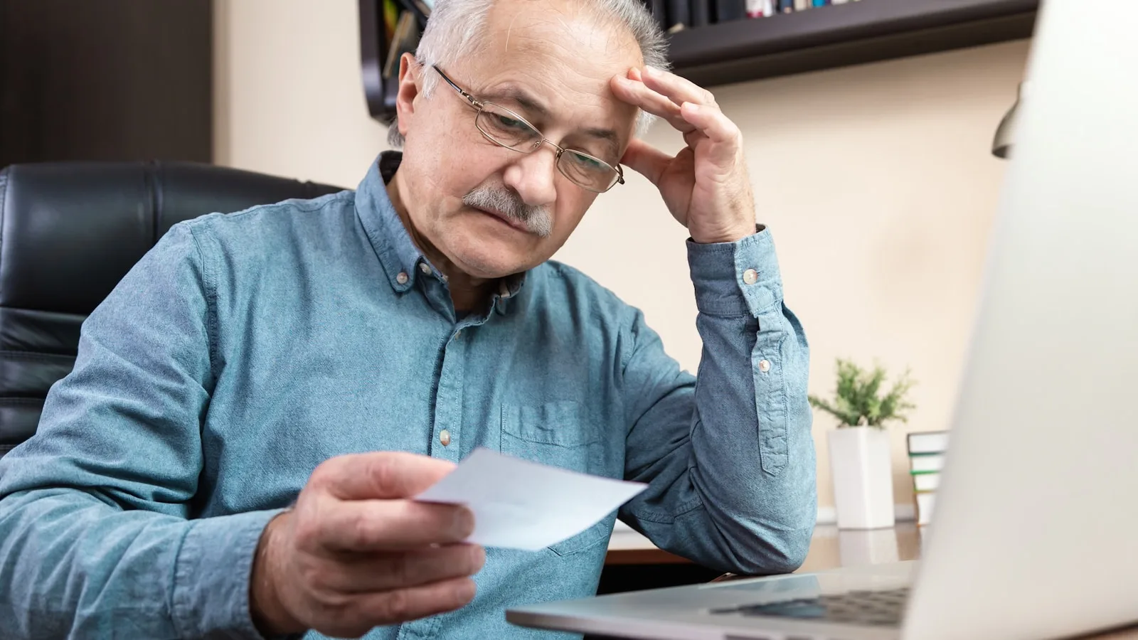 Hombre mayor con gafas y camisa azul, mirando un papel con expresión preocupada frente a un portátil.