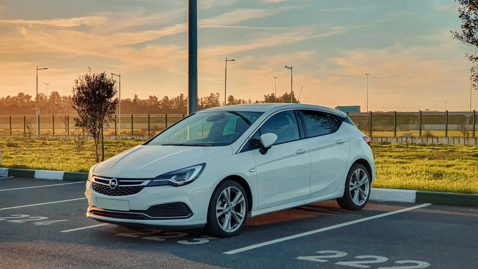 Coche blanco Opel Astra estacionado en plaza numerada al atardecer, con césped y valla al fondo.