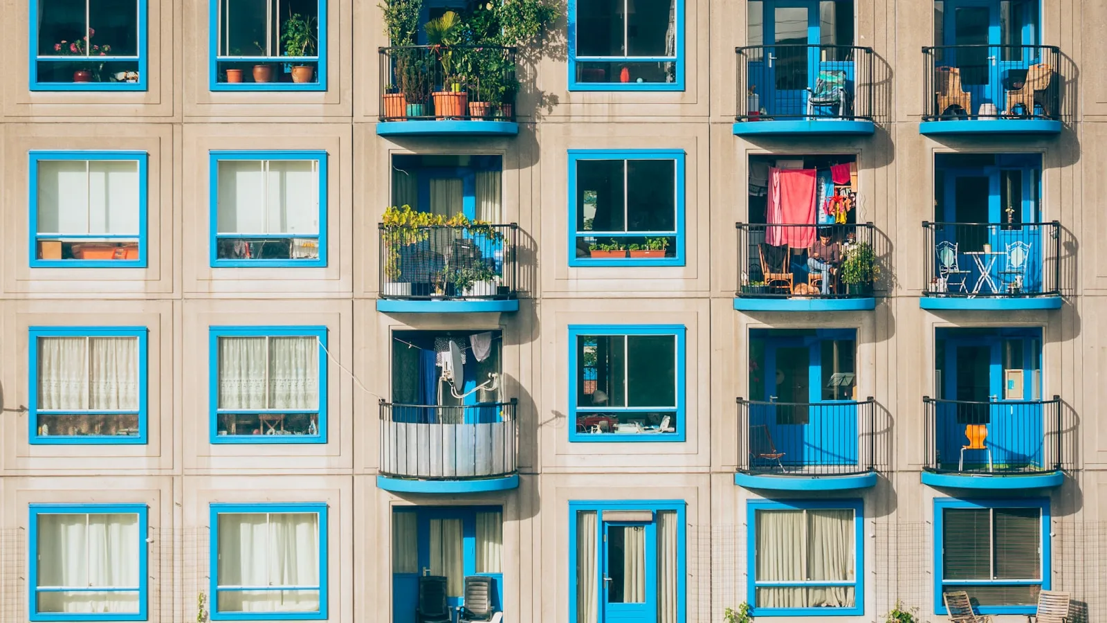 Fachada de edificio residencial con ventanas y balcones de marco azul intenso, algunos con plantas, ropa tendida y muebles.