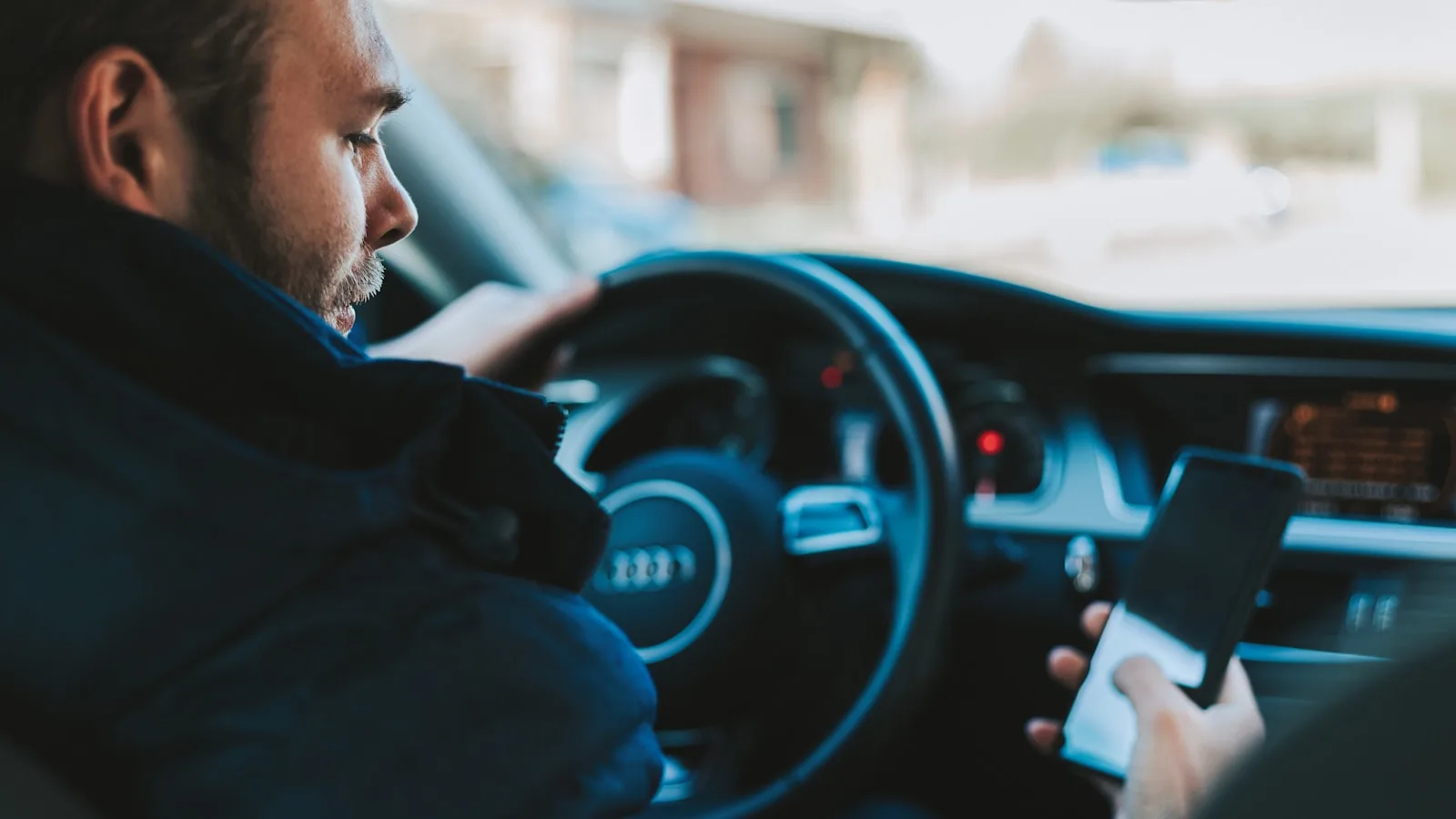 Hombre al volante de un coche (Audi) mirando su teléfono móvil mientras conduce.