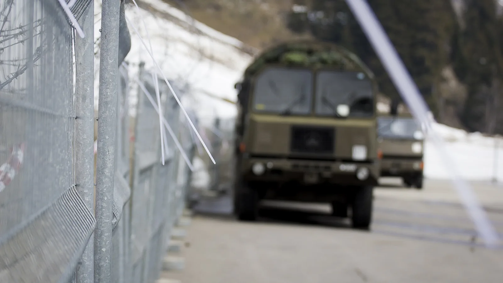 Valla metálica con alambre de espino en primer plano y camión militar desenfocado al fondo.