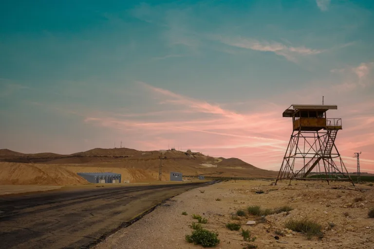 Torre de vigilancia metálica en un paisaje árido al atardecer, con carretera y edificaciones simples al fondo.