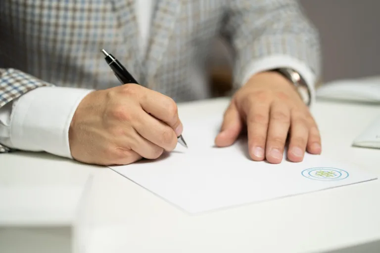 Mano derecha escribiendo con bolígrafo sobre papel blanco; mano izquierda apoyada, con logo circular en la esquina inferior