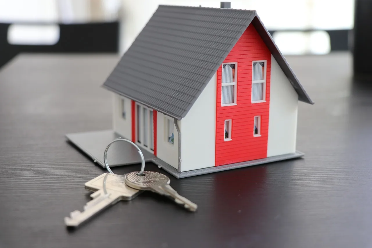 Maqueta de casa en miniatura con techo gris, fachada roja y blanca, y llaves metálicas delante sobre mesa oscura.