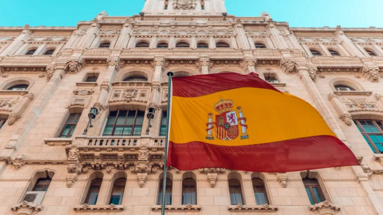Bandera de España ondeando frente a un edificio institucional de arquitectura neoclásica bajo cielo azul claro.