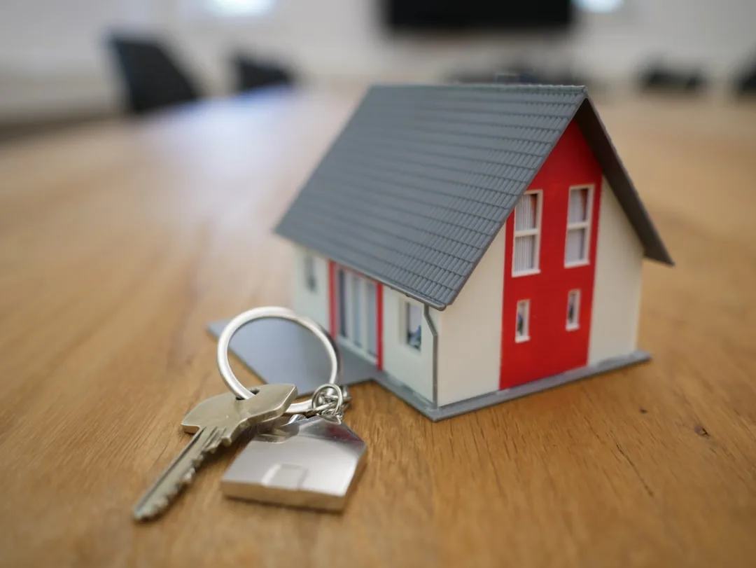 Miniatura de casa de juguete roja y blanca con techo gris, junto a llaves metálicas sobre mesa de madera.